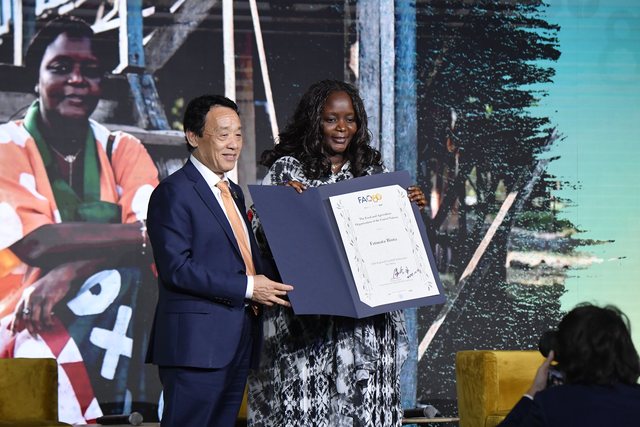Chef Binta on stage receiving an open certificate from a man in a suit, formal backdrop.