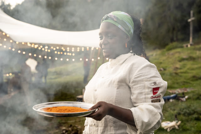 Chef Binta carrying a plate at dusk, string lights overhead.