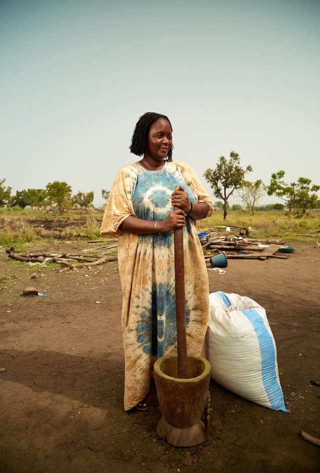 Chef Binta standing outdoors in a flowing tie-dye robe, holding a tall wooden pestle upright, grass and trees behind her.