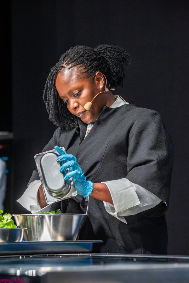 Chef Binta on stage with a headset microphone, mid-demonstration, pouring from a small vessel.