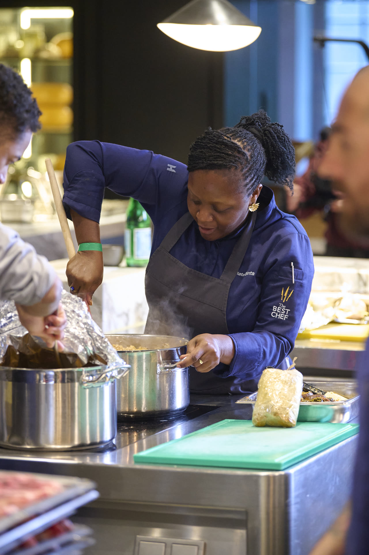 Chef Binta stirring a pot on the stove while another cook assists beside her.