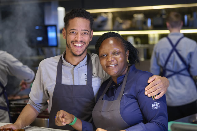 Chef Binta and a teammate embracing at the kitchen pass, both smiling.