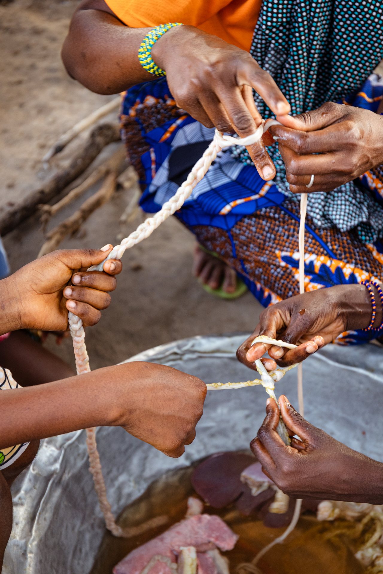 Close on hands braiding pale cord together, seated on the ground.