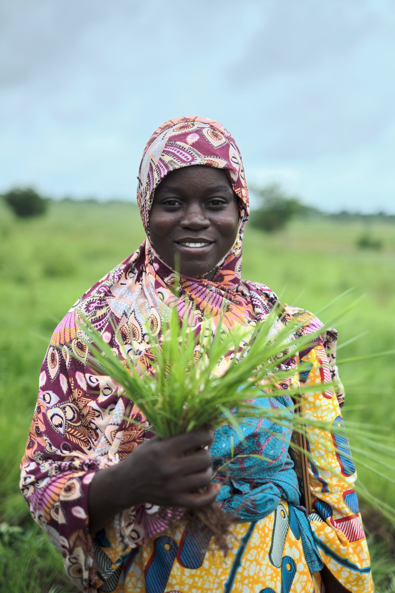 A young woman in a patterned headscarf stands in a green field, holding freshly picked fonio stalks up to the camera.