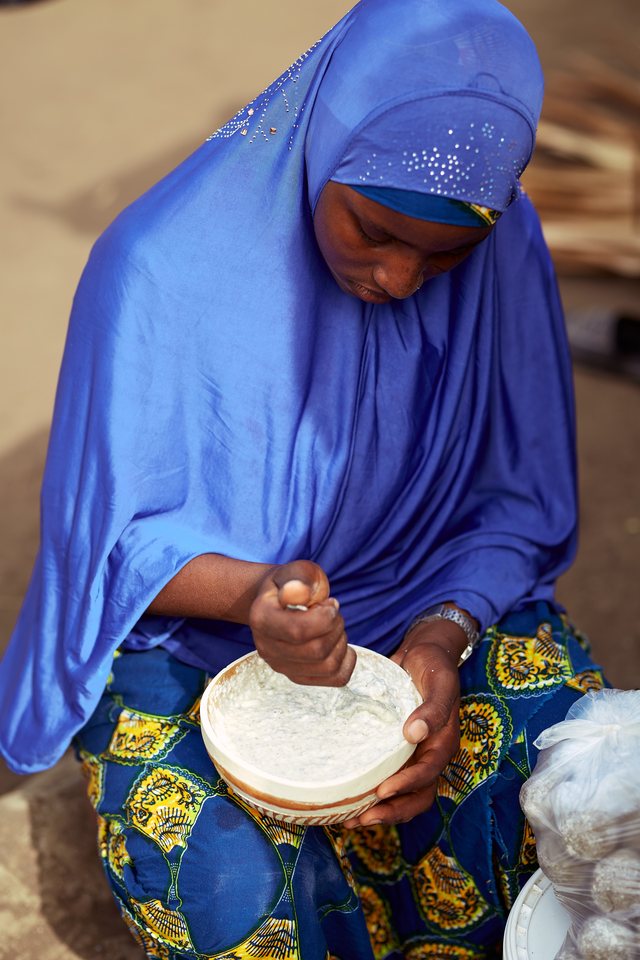 A woman in a blue hijab grinds fonio by hand in a white bowl, working it with her knuckles.