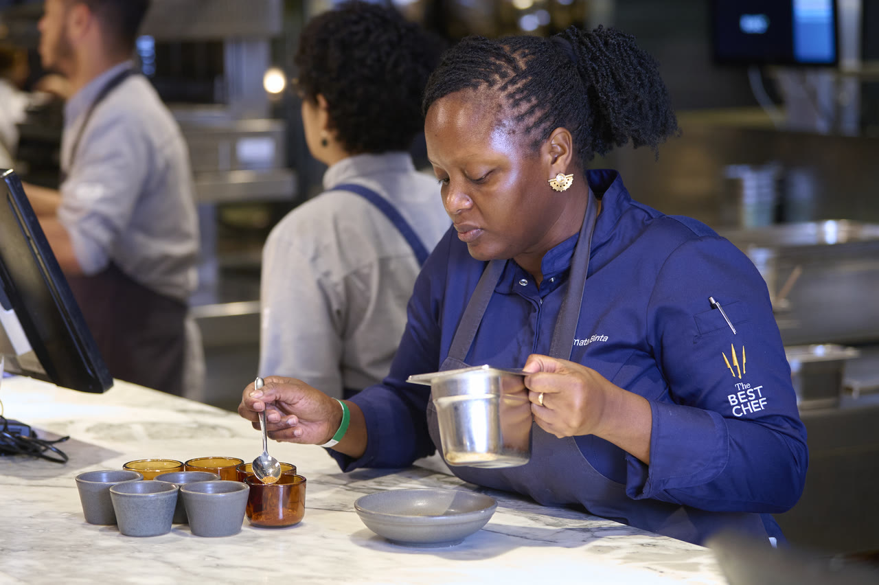 Chef Binta and her team, in blue aprons, prepping dishes at a kitchen pass.