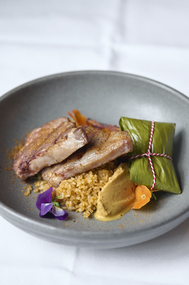 Looking down at a plated dish: sliced meat, a leaf-wrapped parcel tied with string, and a mound of grain, on a pale marble table.