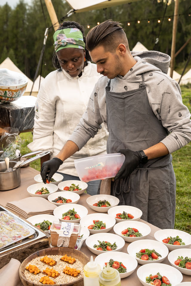 Two cooks plating a row of dishes at an outdoor table beside a tent.