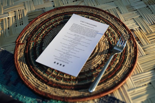Looking straight down at a place setting: a woven charger plate, a folded napkin, cutlery and a printed menu card.