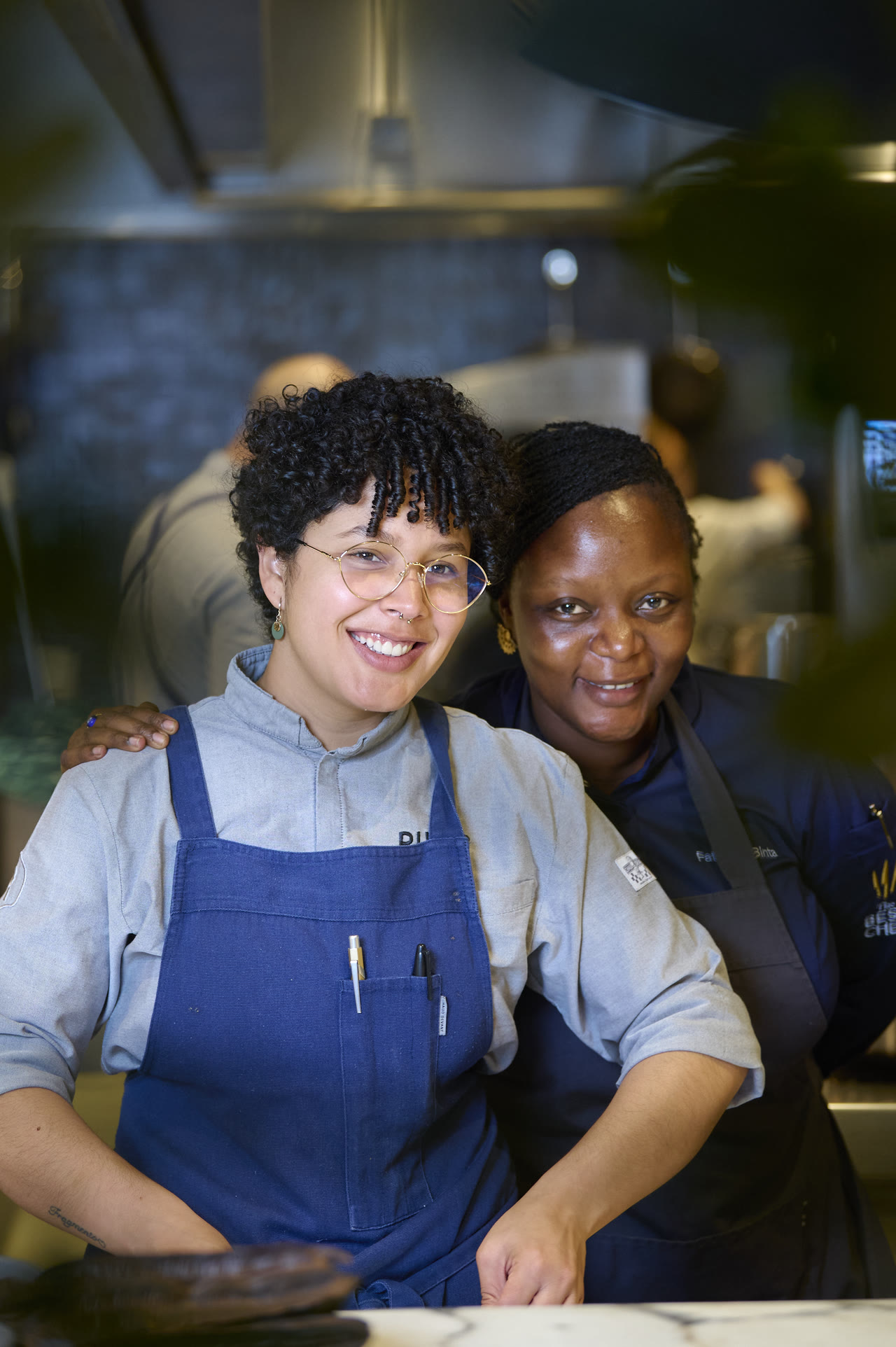 Two women in blue aprons embracing in the kitchen, both smiling.