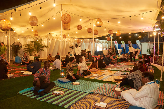 A wide view inside a lit tent: guests seated on patterned mats for dinner, string lights overhead.