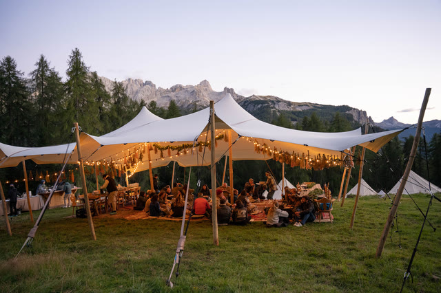 A large white stretch tent set up in a grassy field at dusk, mountains behind it, string lights lit.