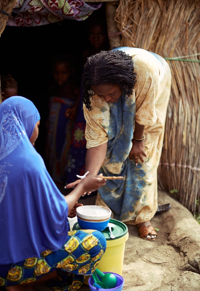 A woman in blue tie-dye bends down to assist an elder woman in a blue hijab.