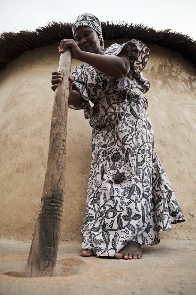A woman pounds grain with a tall wooden pestle against the wall of a mud-brick building.